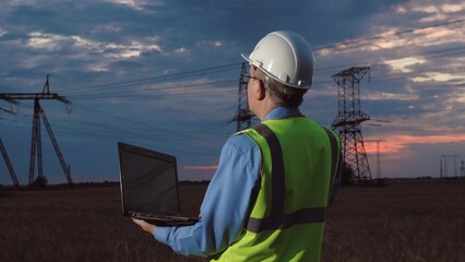 Safety inspector with laptop looks around controlling powerlines network in sunset field. Mature...