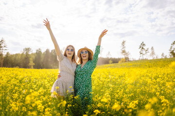 Naklejka na ściany i meble Two young happy women in the middle of a yellow rapeseed field. Girlfriends having fun outdoors on a sunny day. Concept of fun, relaxation. Lifestyle.