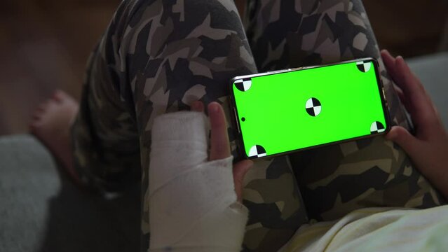 Child with fracture holds phone with chromakey screen in horizontal orientation. Concept of watching online video or making video call on smartphone via Internet by boy wearing cast on his arm.