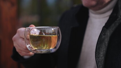 Smiling man drinks light tea sitting in cafe gazebo looking to side. Tea drinking arranged by man to meet friend in cafe gazebo. Man taking break from work and drinking tea in cafe gazebo