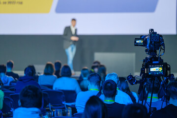 Video camera recording male speaker giving presentation in illuminated conference hall