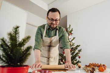 Happy man in glasses wearing apron and rolling out the dough for christmas cookies