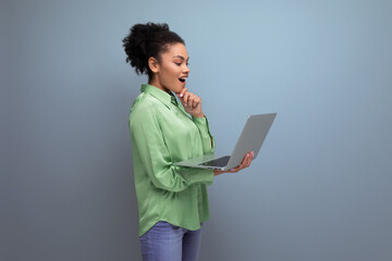 young brunette hispanic woman with curly hair in green rejoices holding a laptop in her hand against the background with copy space