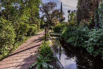 A stream of water next to a red cobblestone path within a plot with gardens and trees