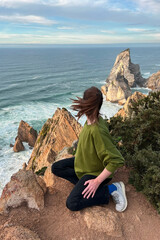 Fototapeta premium Young woman sitting on the cliff and looking at the Atlantic ocean in Portugal