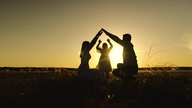 Silhouettes of parents against sky over daughter holding hands with setting sun. Figures of parents above girl jumping under imagined roof. Silhouettes of parents making shape of dream house with arms