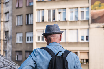 A man with a fedora and a backpack watches the buildings in front of him