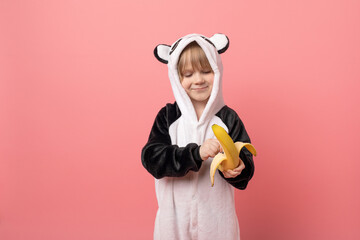 Child in panda animal costume eats banana on pink background, studio shot.