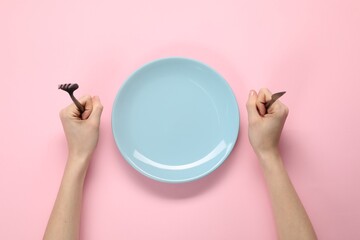 Woman with fork, knife and empty plate at pink table, top view