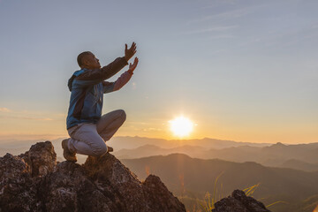Hiker male kneeling down with hands open palm up praying to God on top mountain sunset background.
