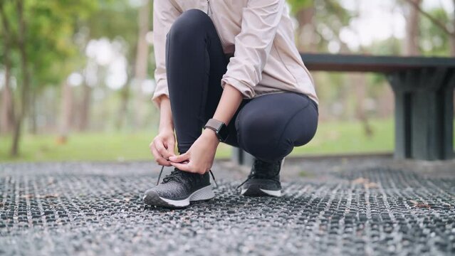 An Asian woman in workout attire is lacing up her shoes, getting ready for exercise in a public park. Capture the preparation and determination for a fitness session outdoors