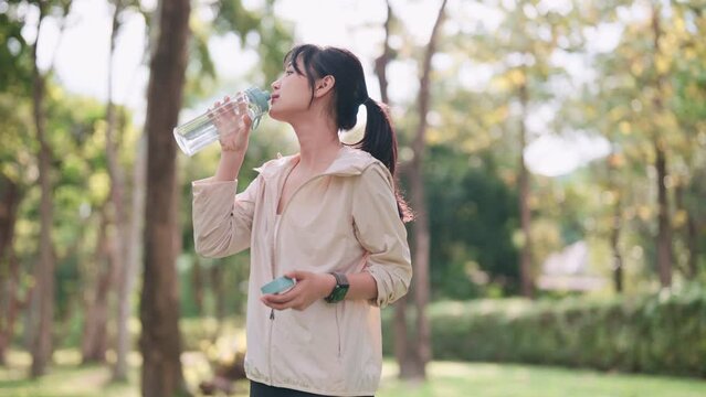 An Asian woman in workout attire is drinking water, taking a break from exercising in a public park. Showcase hydration and wellness during a fitness routine outdoors for a refreshing image