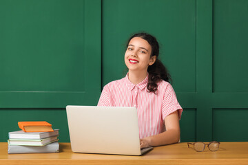 Female student with laptop doing homework at table near green wall