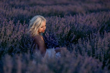 Fototapeta na wymiar Blonde woman poses in lavender field at sunset. Happy woman in white dress holds lavender bouquet. Aromatherapy concept, lavender oil, photo session in lavender
