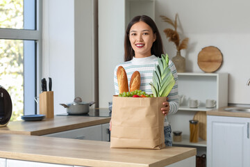 Young Asian woman with shopping bag full of fresh food at table in kitchen