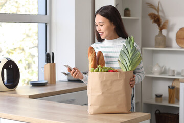 Young Asian woman with mobile phone and shopping bag full of fresh food at table in kitchen