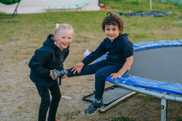 Fototapeta premium Two children playing outside, a girl with a flower in her hair laughing and a boy in a hoodie sitting on the edge of a trampoline.