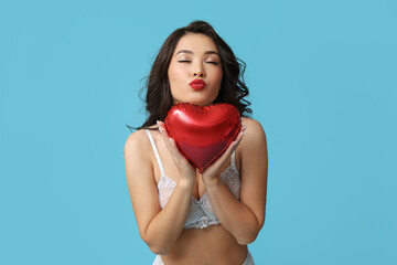 Beautiful young Asian woman in sexy underwear with heart-shaped balloon blowing kiss on blue background