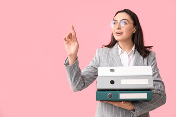 Young businesswoman in eyeglasses with document folders pointing at something on pink background
