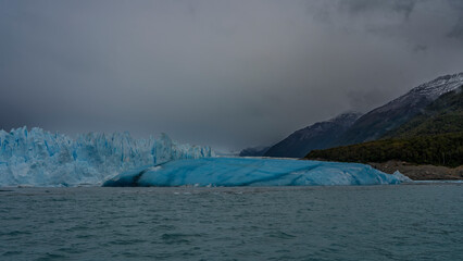 The beautiful glacier of Patagonia. A wall of cracked blue ice in a turquoise lake. A large breakaway iceberg floats in the water. Coastal mountains in clouds and fog. Perito Moreno. El Calafate. 