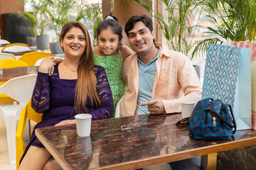 Happy indian family of three sitting in shopping mall cafeteria enjoying with cup of coffee and...