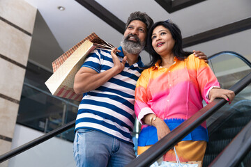 Happy indian couple holding on escalator handrail and riding escalator in shopping mall with paper...