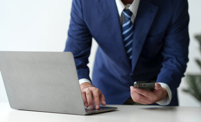 Businessman holds smartphone, checks email, checks job details on Laptop computer on white desk to...
