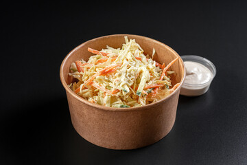 Vitamin salad with fresh cabbage in paper take away container with sauce on black background.