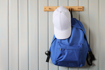 Blue backpacks and a white baseball cap are hanging on a wooden hanger on a board wall.