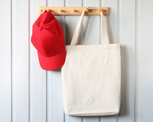 white cloth bag and a baseball cap hang on a wooden hanger on a white board wall. mockup, creator...