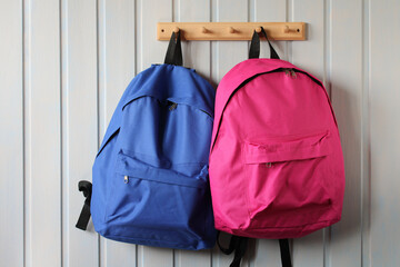 blue and pink school backpacks hang on a wooden hanger on a white board wall.