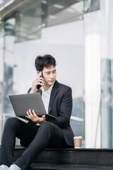 Smiling Asian young freelancer working outside an office building remotely. He is talking on the phone.