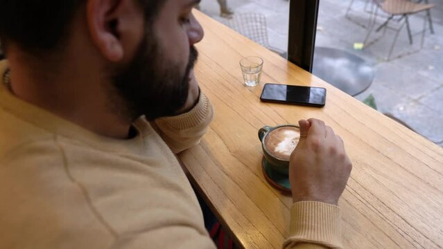 A bearded guy is stirring the coffee while feeling loneliness near the window. Copy space.