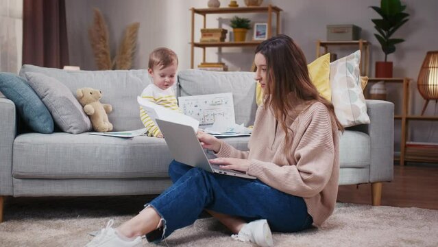 Woman sitting at floor carpet and holding laptop. Mother working online and taking care of daughter. Baby girl sitting at sofa and playing with business papers. Mother and toddler at home.