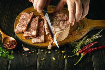 The cook cuts the brawn into pieces with a knife on the kitchen board before preparing delicious...