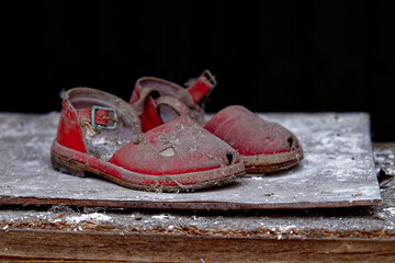 Two worn-out red sandals with buckles, lying on a dusty and dirty surface.