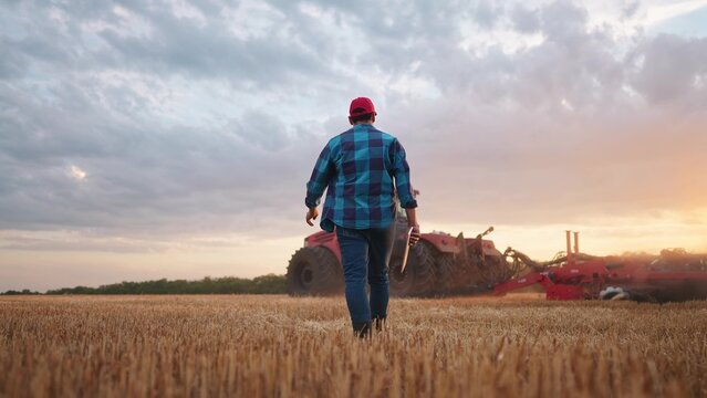agriculture. farmer walk works in a field next to a tractor that plows the land. business agriculture concept. farmer with tablet works in a field next to a tractor at farm sunset mowed wheat plows
