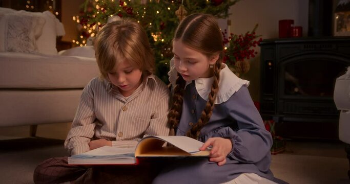 blonde boy and girl with two braids have fun near Christmas tree at home, sit on floor reading a book together and looking at pictures, talking, whispering, smiling and laughing