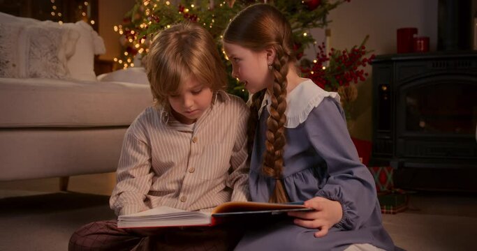 blonde boy and girl with two braids have fun near Christmas tree at home, sit on floor reading a book together and looking at pictures, talking, whispering, smiling and laughing