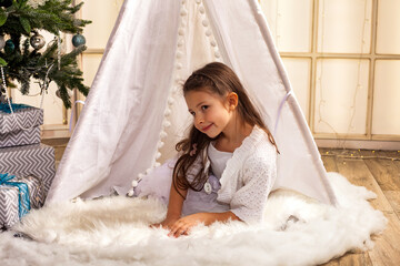 Smiling child girl in white dress posing in wigwam at Christmas tree, happy looking at camera. Cover kid girl 5 year old lying down in children room. Christmas magical concept. Copy ad text space