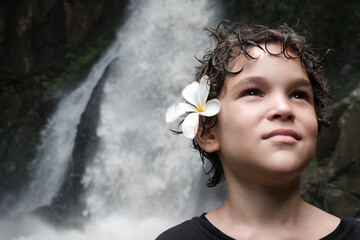 Portrait of a boy with plumeria (frangipani) flower in his hairs on the background of waterfall. Bali, Indonesia.