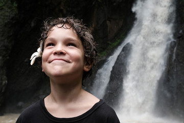 Happy Caucasian boy with plumeria (frangipani) flower in his hairs smiles on the background of Singsing waterfall. Bali, Indonesia.