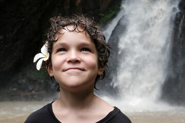 Portrait of a Caucasian boy with plumeria (frangipani) flower in his hairs smiling on the background of waterfall. Bali, Indonesia.