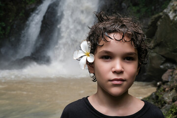 Portrait of a Caucasian boy with plumeria (frangipani) flower in his hairs on the background of Singsing waterfall. Bali, Indonesia.