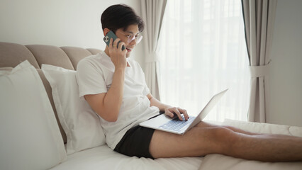 Businessman in wears casual clothes talking on mobile phone while using laptop on bed. He enjoys...