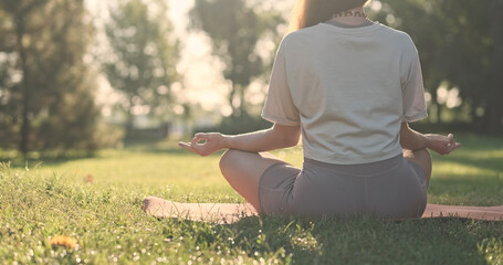 Yoga mudra, woman sitting on mat and putting her fingers together, meditation in nature, city park, dawn, green lawn, back view