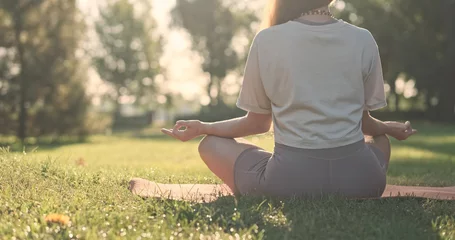 Zelfklevend Fotobehang Yoga mudra, woman sitting on mat and putting her fingers together, meditation in nature, city park, dawn, green lawn, back view © Oleksii