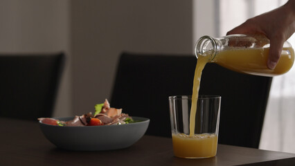 Pour orange juice next to salad with tomatoes, lettuce and prosciutto cotto in blue bowl on wood table