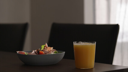 orange juice next to salad with tomatoes, lettuce and prosciutto cotto in blue bowl on wood table