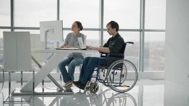 Paralyzed man work with woman in office. Colleagues discussing project together female shows to guy in wheelchair pie chart on paper document. Positive workers discuss analytics data sit at desk.